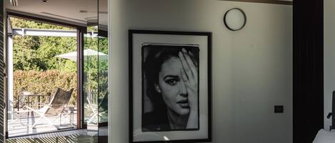 A modern hotel room with a large bed, a framed portrait on the wall, and a view of the outdoors through a glass door.