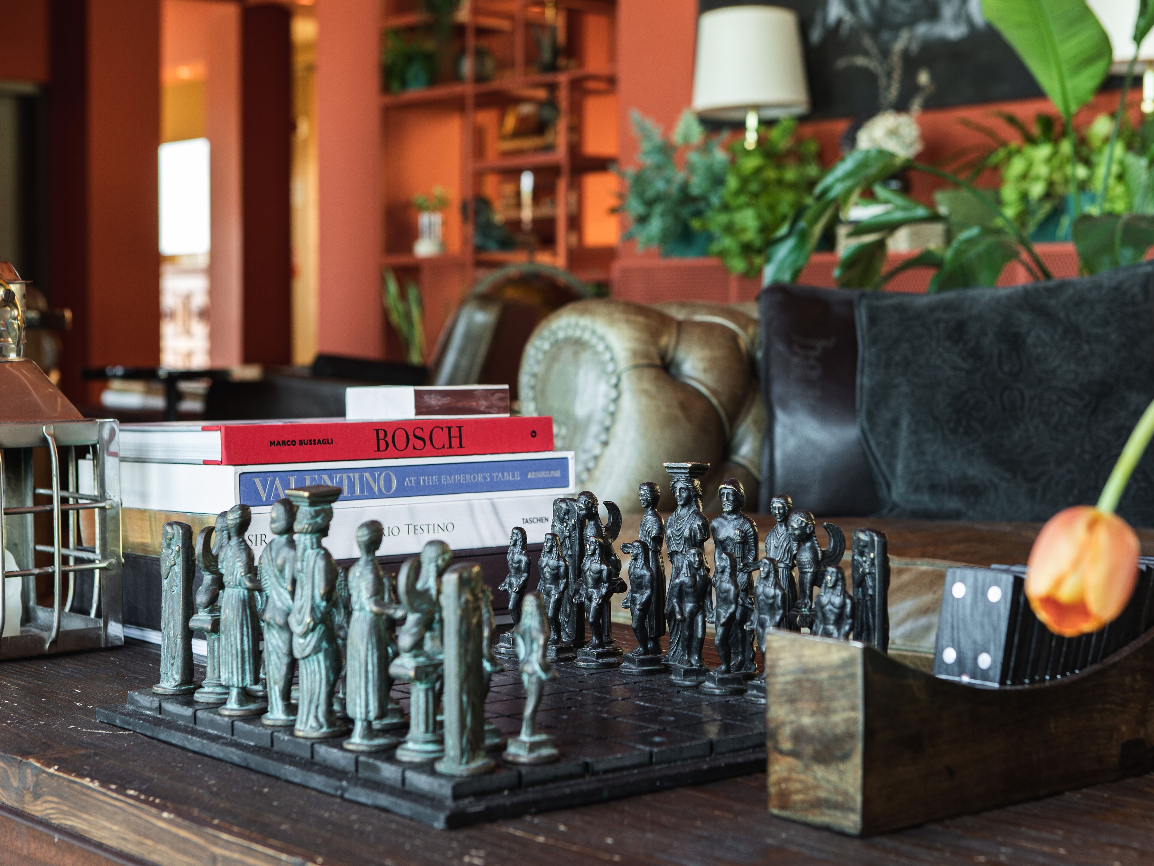 A chessboard with figurines, a stack of books, and a leather couch in a room with a bookshelf and a lamp.