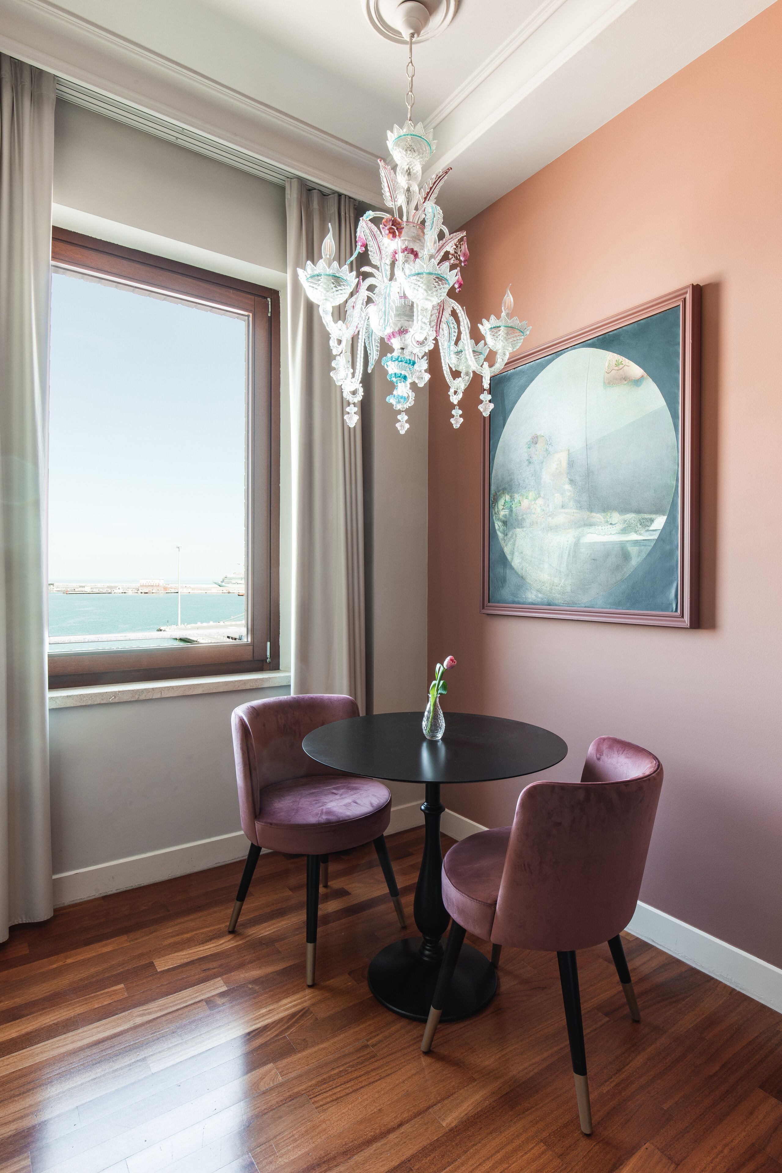 A dining area with a round table, two chairs, a chandelier, a window with curtains, and a framed artwork on the wall.