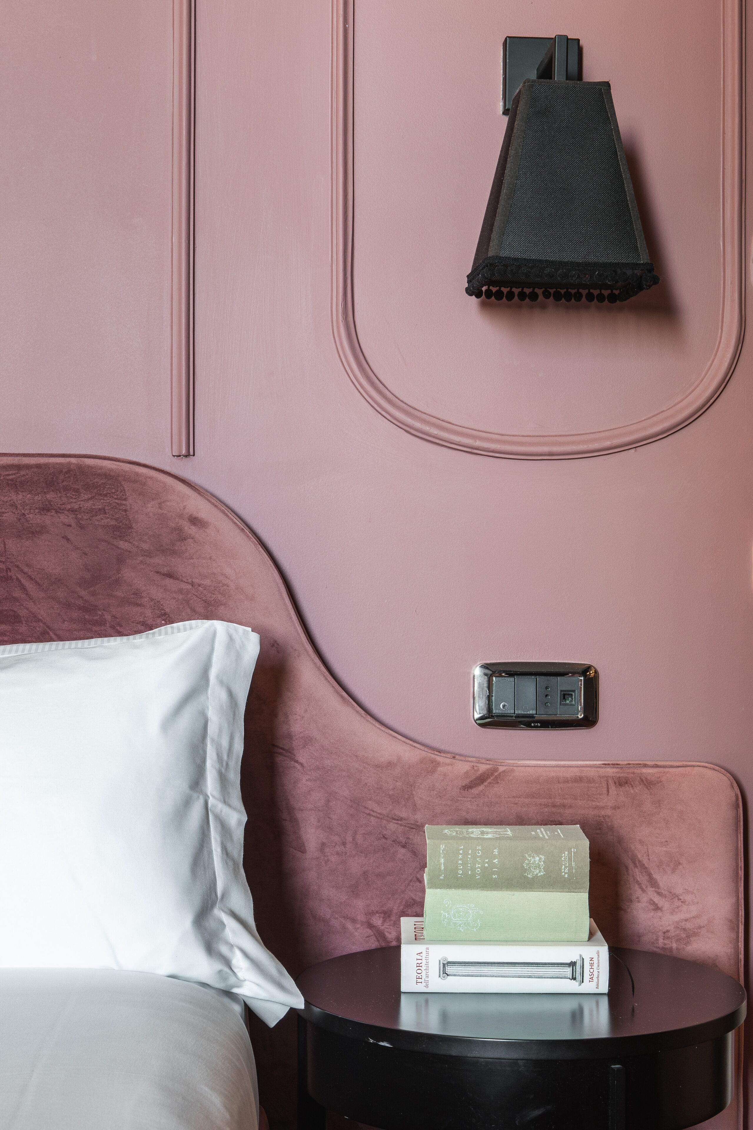 A bed with a white pillow and a pink headboard, a bedside table with books, and a wall-mounted lamp.