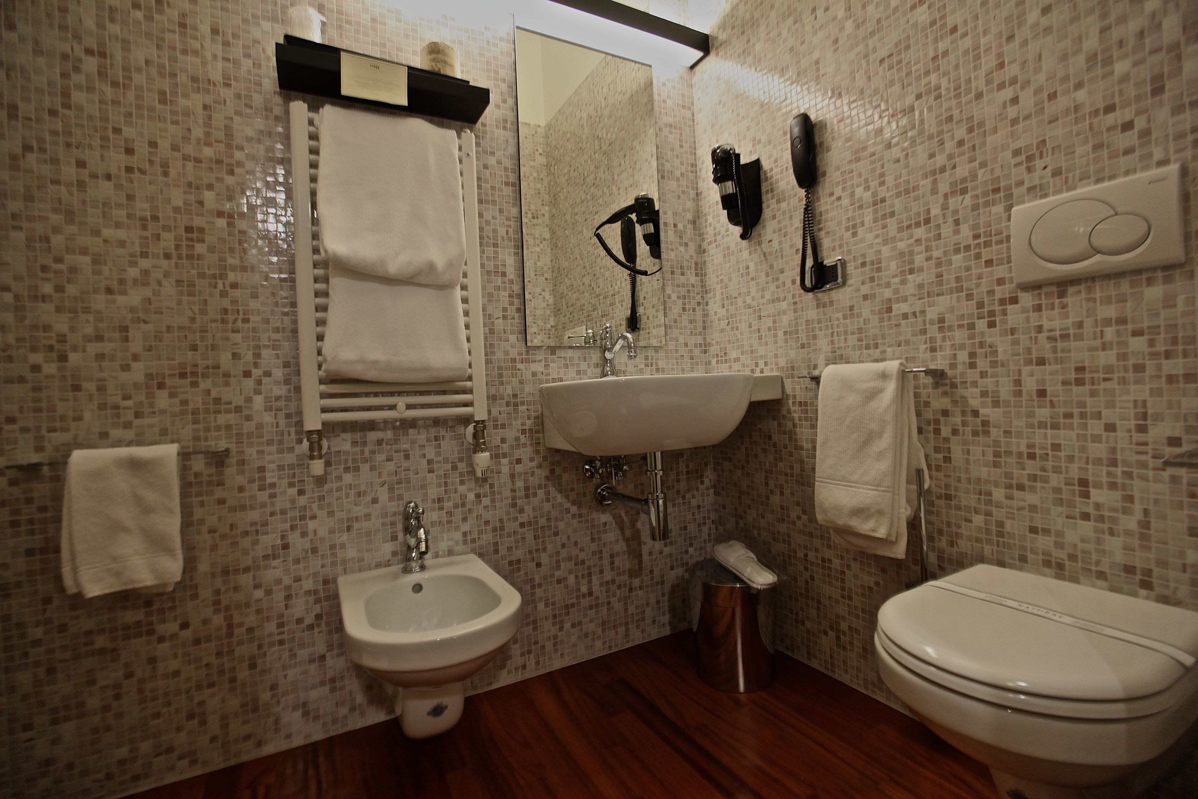 A bathroom with a mosaic tile wall, a sink, a bidet, a toilet, a mirror, and a hairdryer.