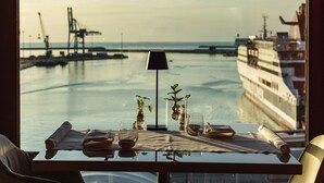 A table with a glass, a vase with a plant, and a rolled-up menu, overlooking a calm sea with a cruise ship and cranes in the background.