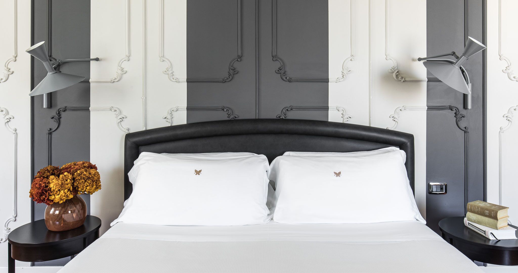 A neatly made bed with white linens and two pillows, flanked by two bedside tables with lamps, a vase of flowers, and a stack of books on one table.