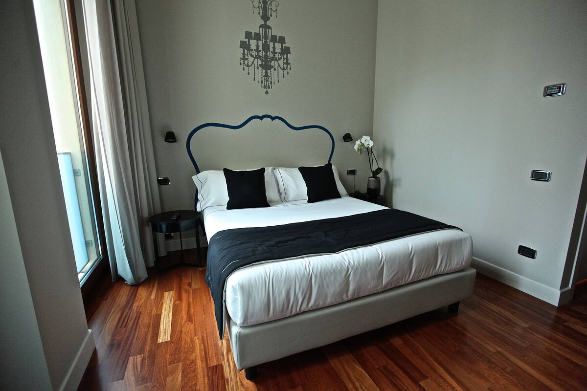 A bedroom with a bed, wooden flooring, a chandelier, and a nightstand with a vase of flowers.