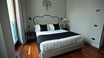 A bedroom with a bed, wooden flooring, a chandelier, and a nightstand with a vase of flowers.