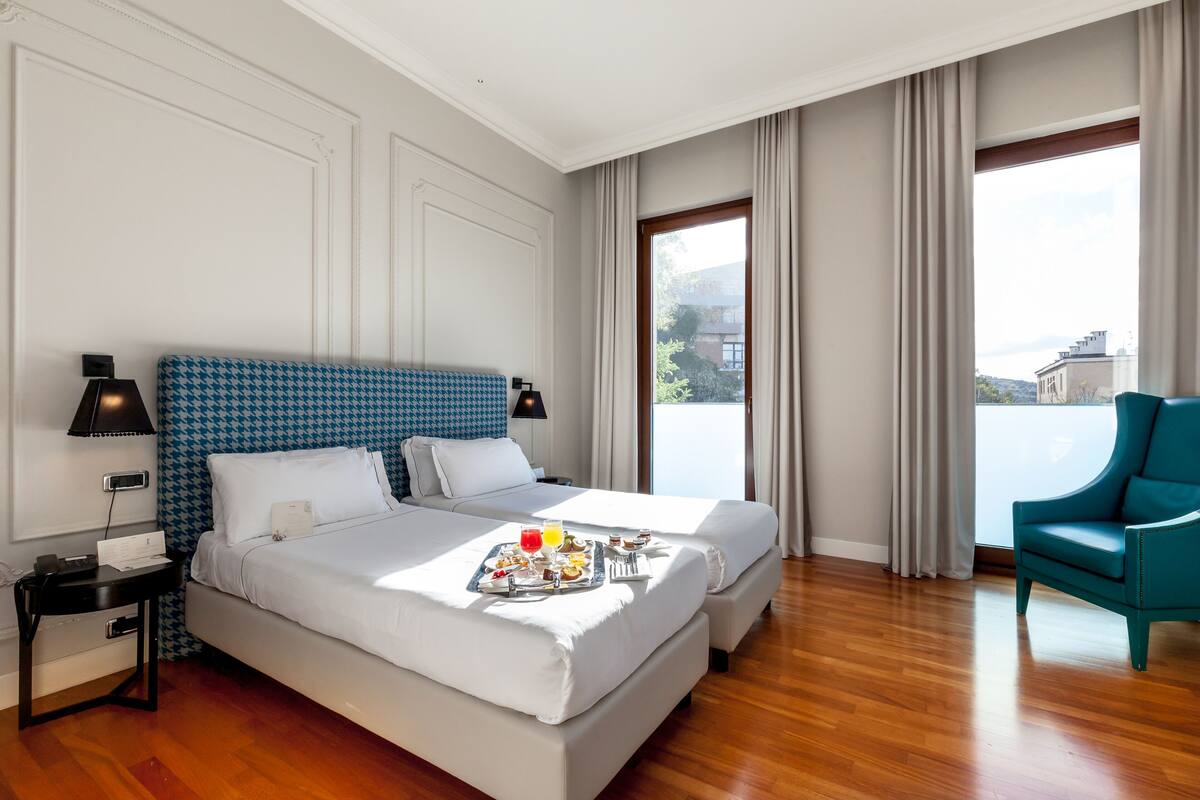A hotel room with two beds, a dining table with breakfast, a chair, and a large window with curtains.