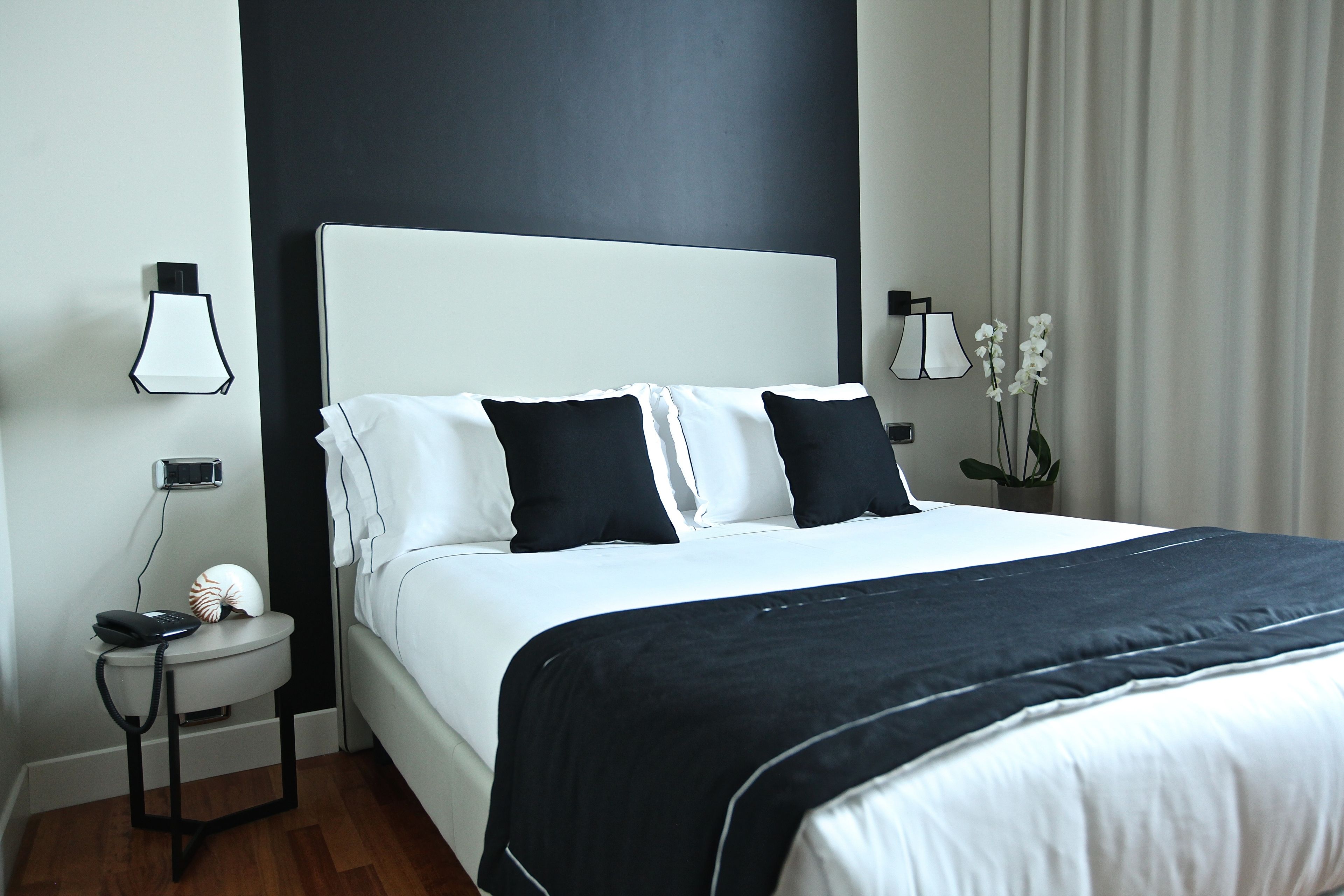 A modern hotel room with a large bed, two bedside tables, a wall-mounted lamp, and a potted orchid plant.