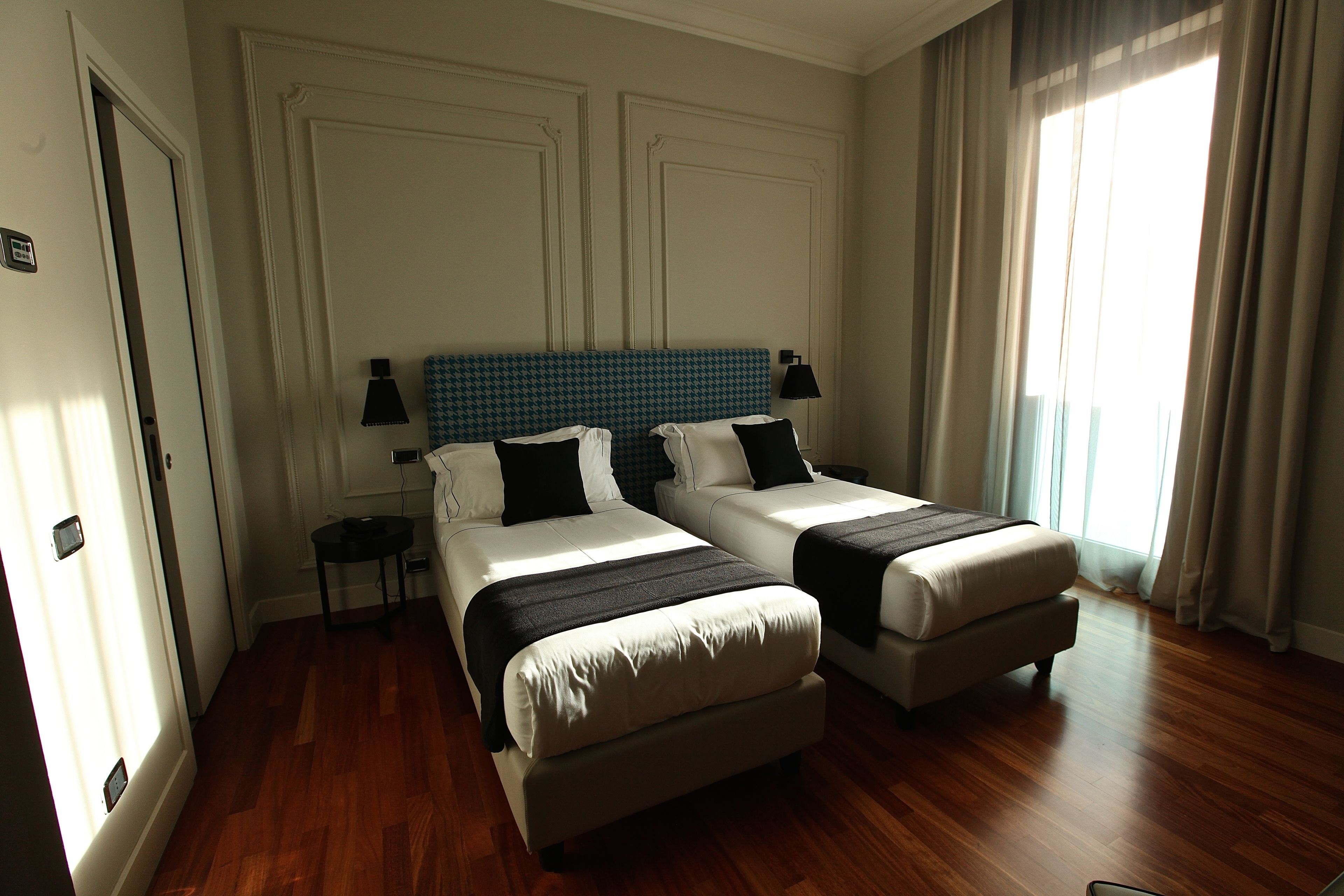 Habitación de hotel con dos camas, suelo de madera, un ventanal grande con cortinas y una pared blanca con un panel decorativo.