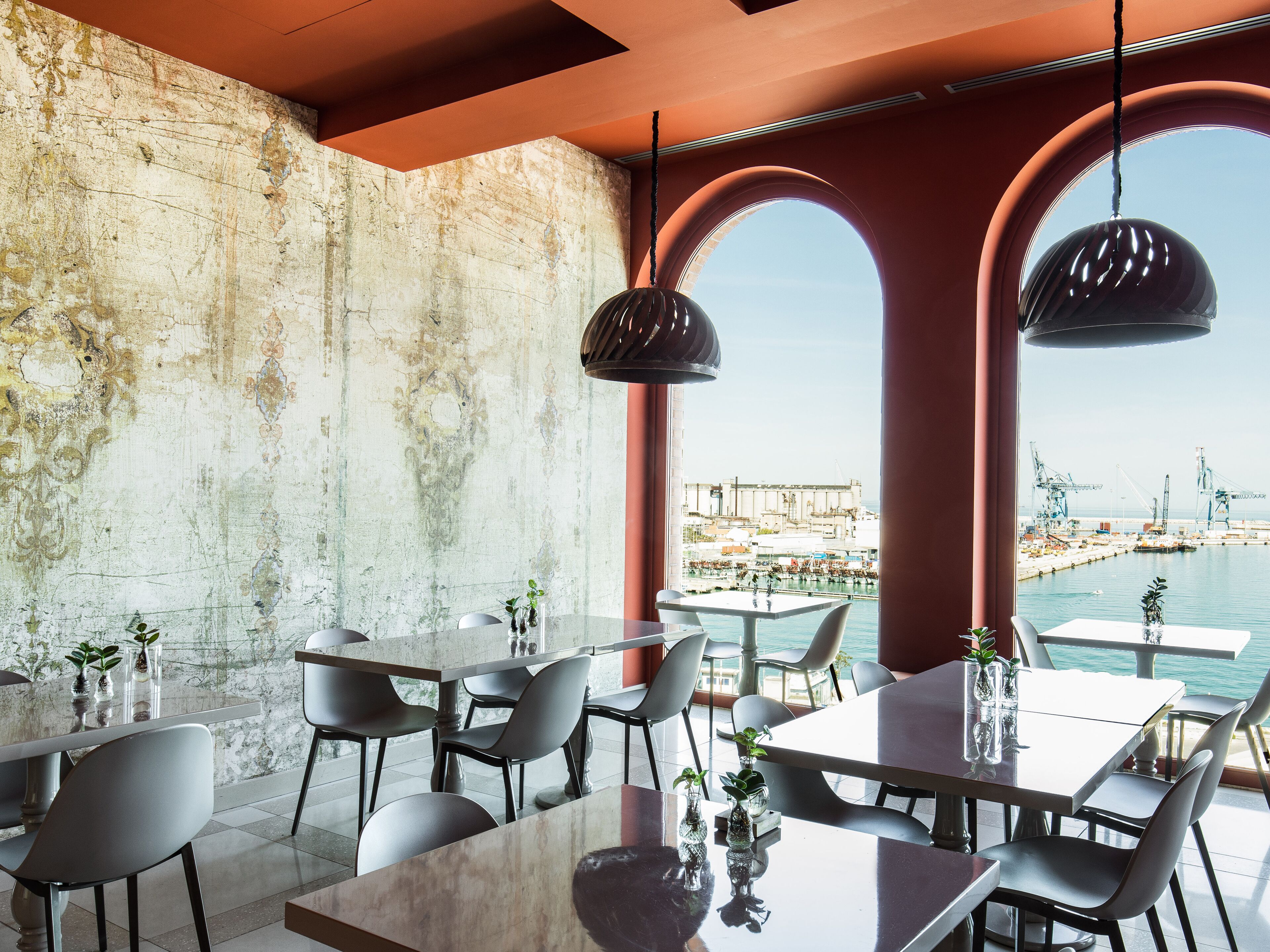 A modern dining area with tables and chairs, a concrete wall, and a view of a port with cranes.
