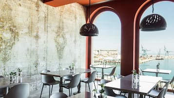 A modern dining area with tables and chairs, a concrete wall, and a view of a port with cranes.