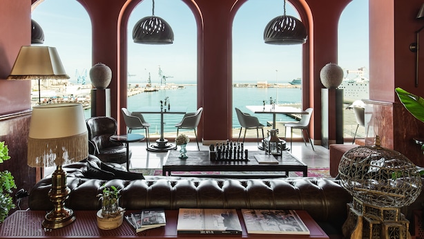 A room with a view of the harbor, featuring a leather sofa, two tables, and a chandelier.