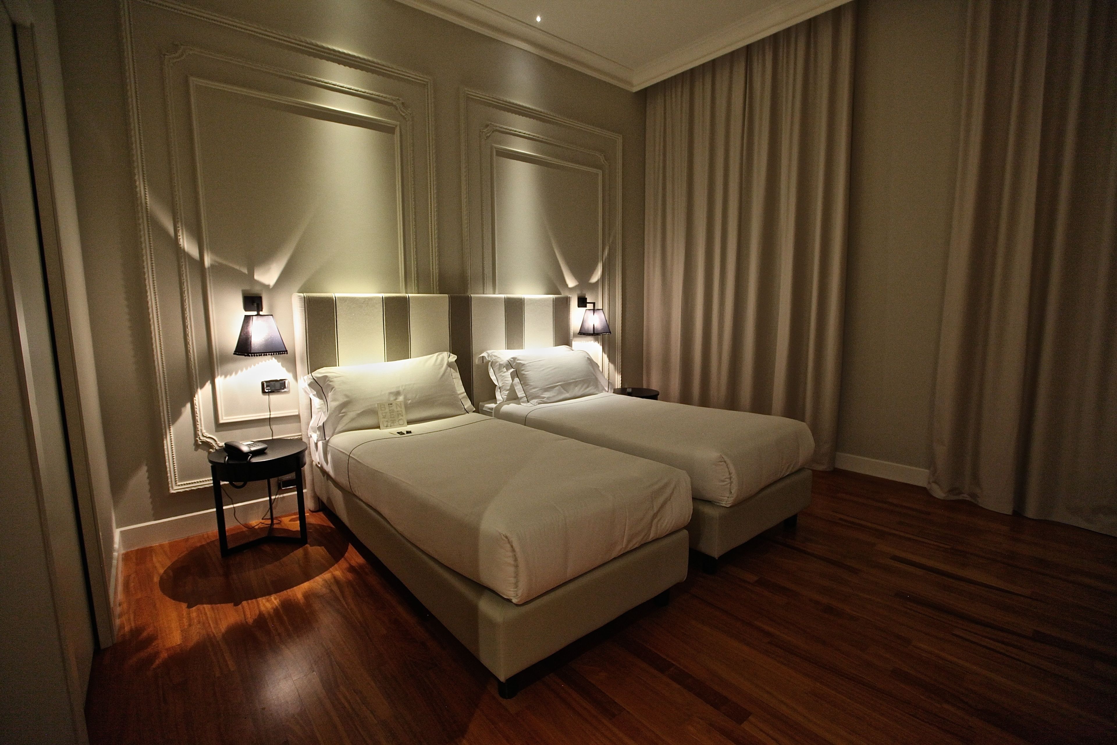 Habitación de hotel con dos camas, una mesita de noche, una lámpara y suelo de madera.