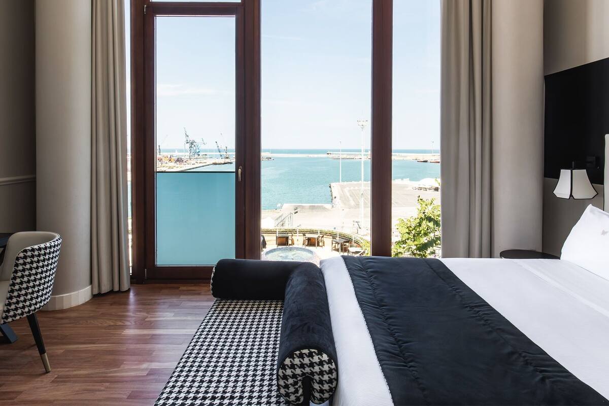 A hotel room with a large bed, a desk with a lamp, a chair, and a view of the sea and port.