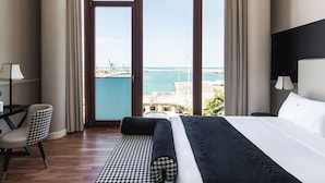 A hotel room with a large bed, a desk with a lamp, a chair, and a view of the sea and port.