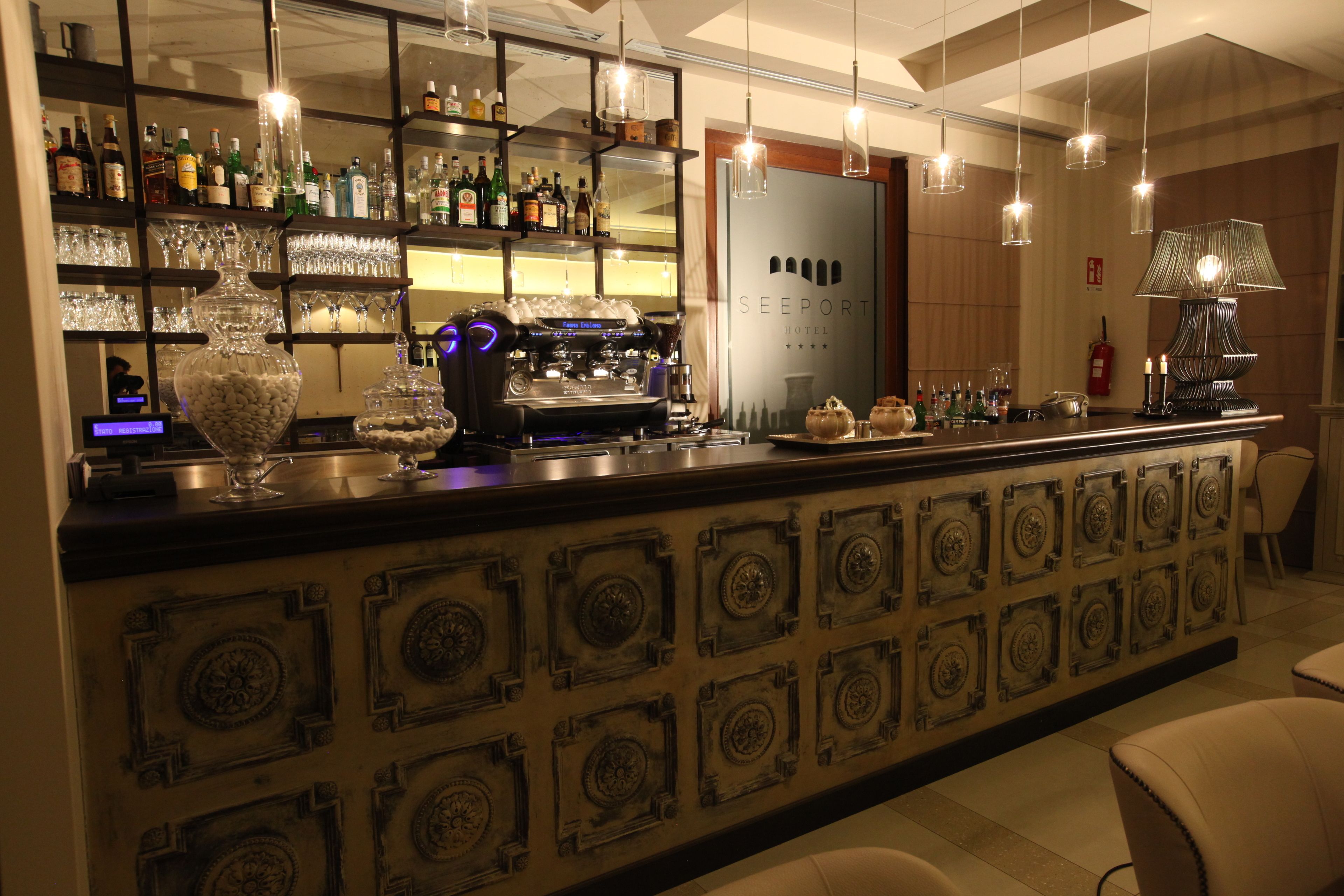 A bar area with shelves of liquor bottles, a coffee machine, and a decorative wall with geometric patterns.