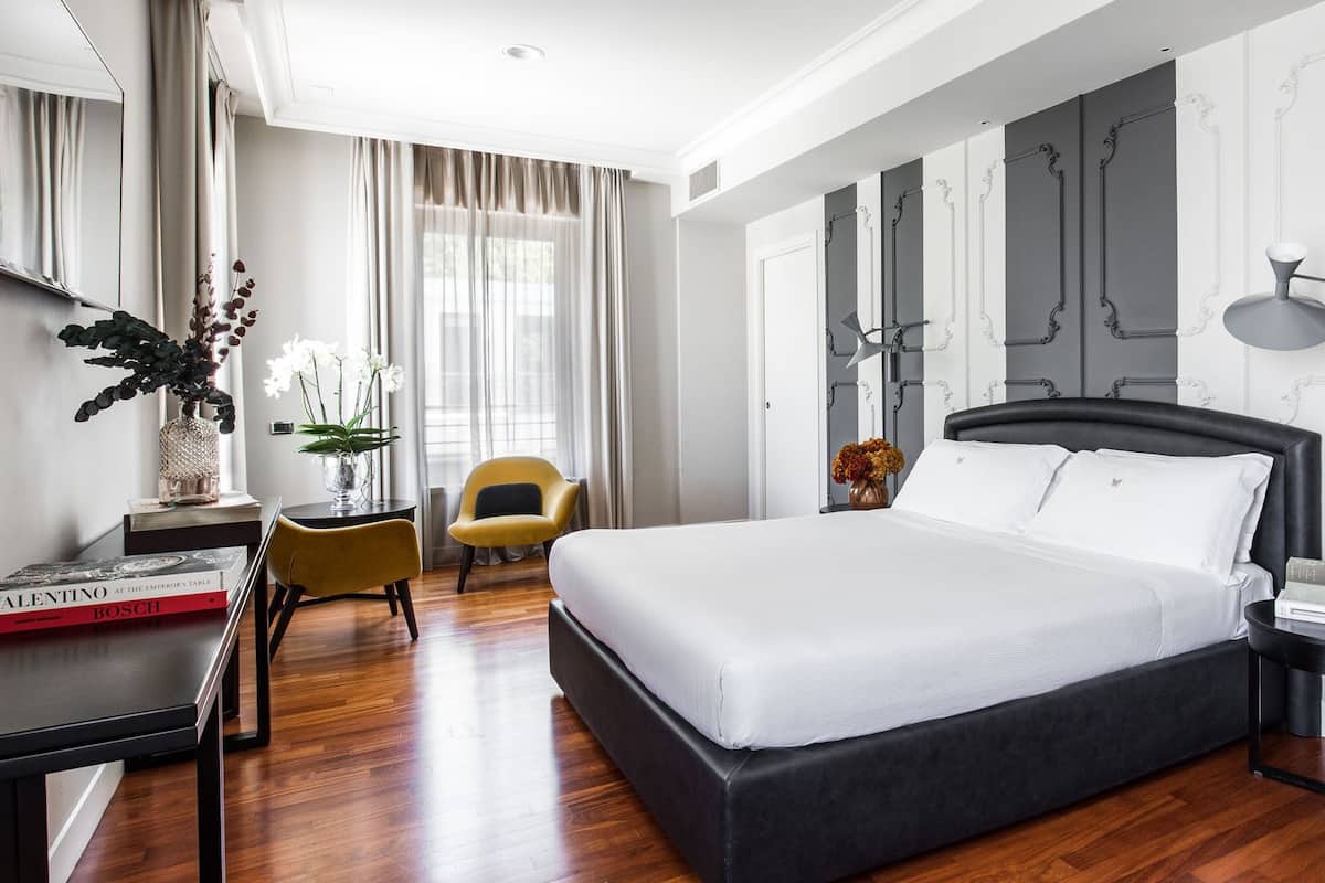 A modern hotel room with a large bed, a desk with a chair, a bedside table, and a window with curtains.