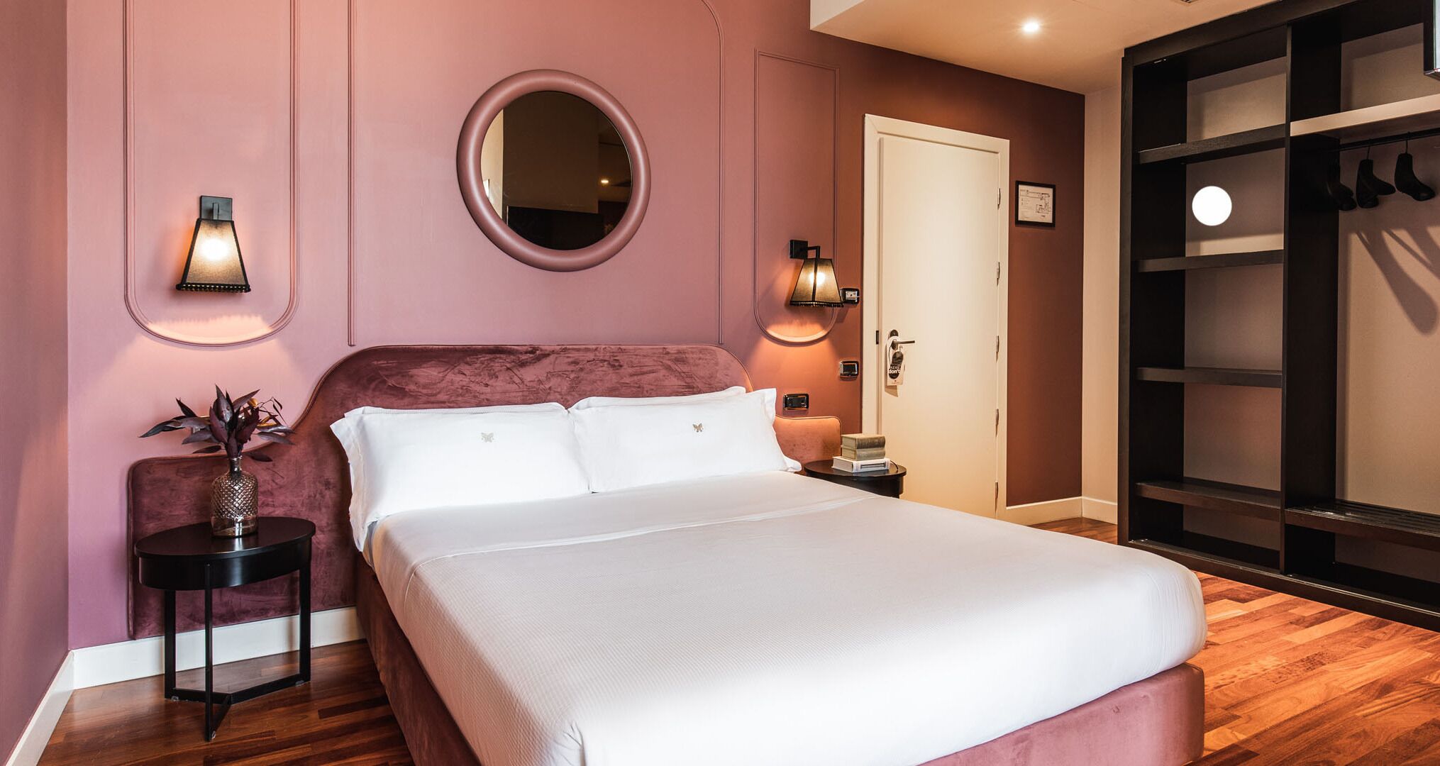 A bedroom with a large bed, a round mirror, two wall sconces, a small side table with a vase, and a wardrobe with hanging shoes.