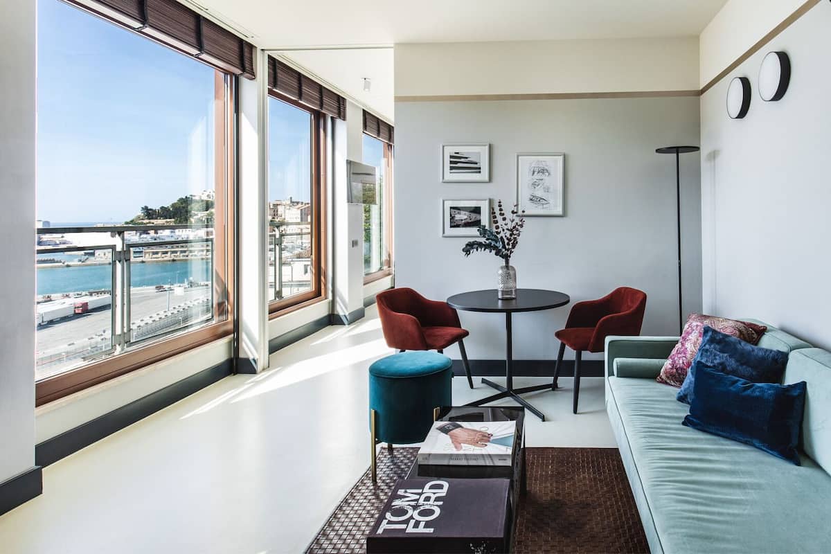 A modern living room with a view of the harbor, a round table with chairs, and a sofa with cushions.