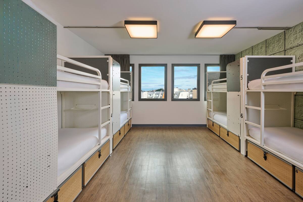 Habitación tipo dormitorio con literas, suelo de madera y ventanas amplias que dan a una vista de la ciudad.
