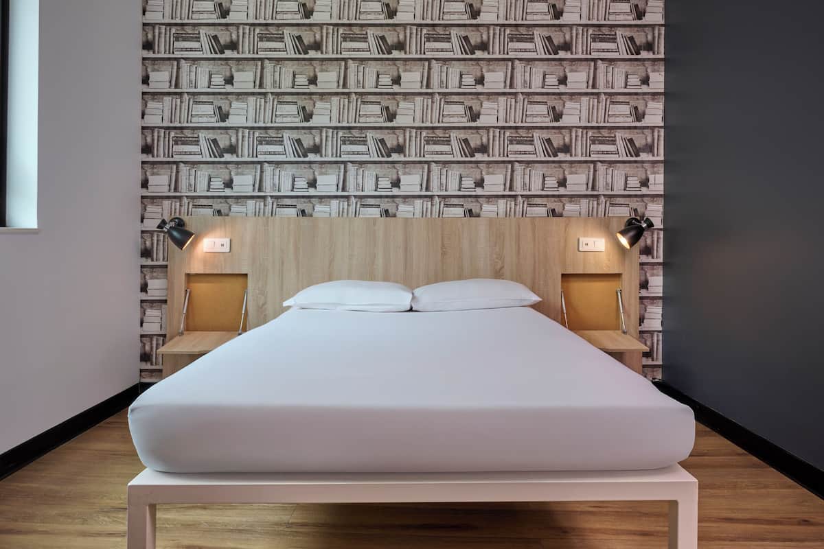 Una cama bien hecha con un colchón y almohadas blancos, sobre una plataforma blanca. La habitación tiene un cabecero de madera y un papel tapiz con diseño de libros.