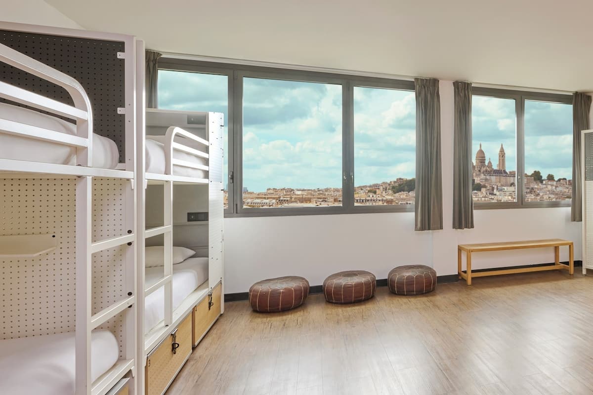 Una habitación con literas, un banco y vistas a un paisaje urbano a través de amplios ventanales.