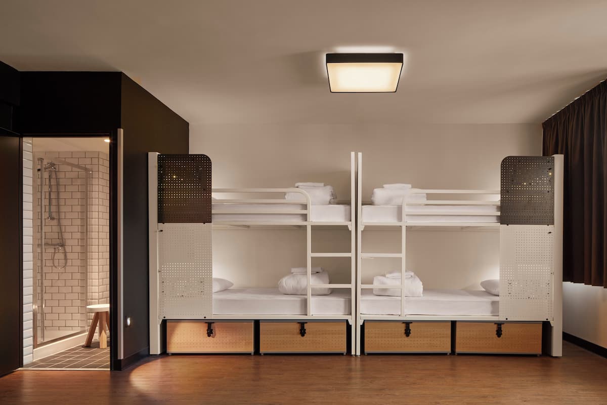 Una habitación de hotel moderna con una cama litera, zona de ducha y un taburete pequeño.
