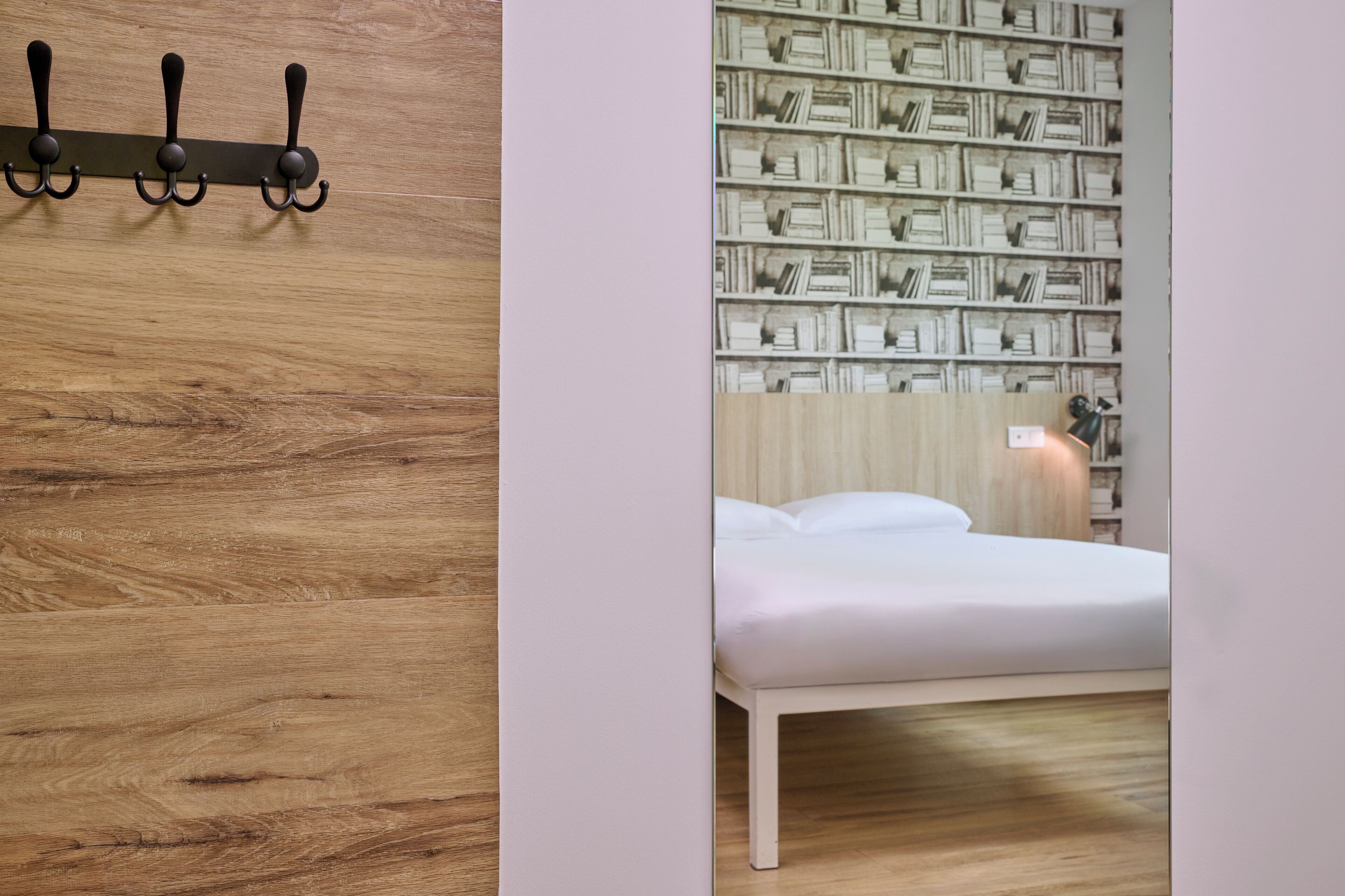 Une chambre avec un plancher en bois, un lit blanc, une bibliothèque fixée au mur et un luminaire fixé au mur.