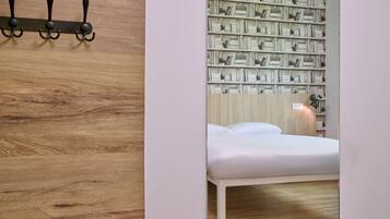 Une chambre avec un plancher en bois, un lit blanc, une bibliothèque fixée au mur et un luminaire fixé au mur.