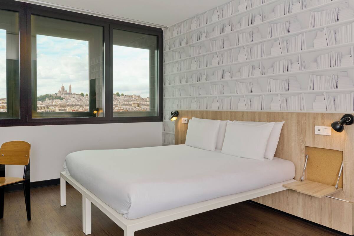 Un dormitorio ordenado con un ventanal grande, una cama, una silla y un estante en la pared.