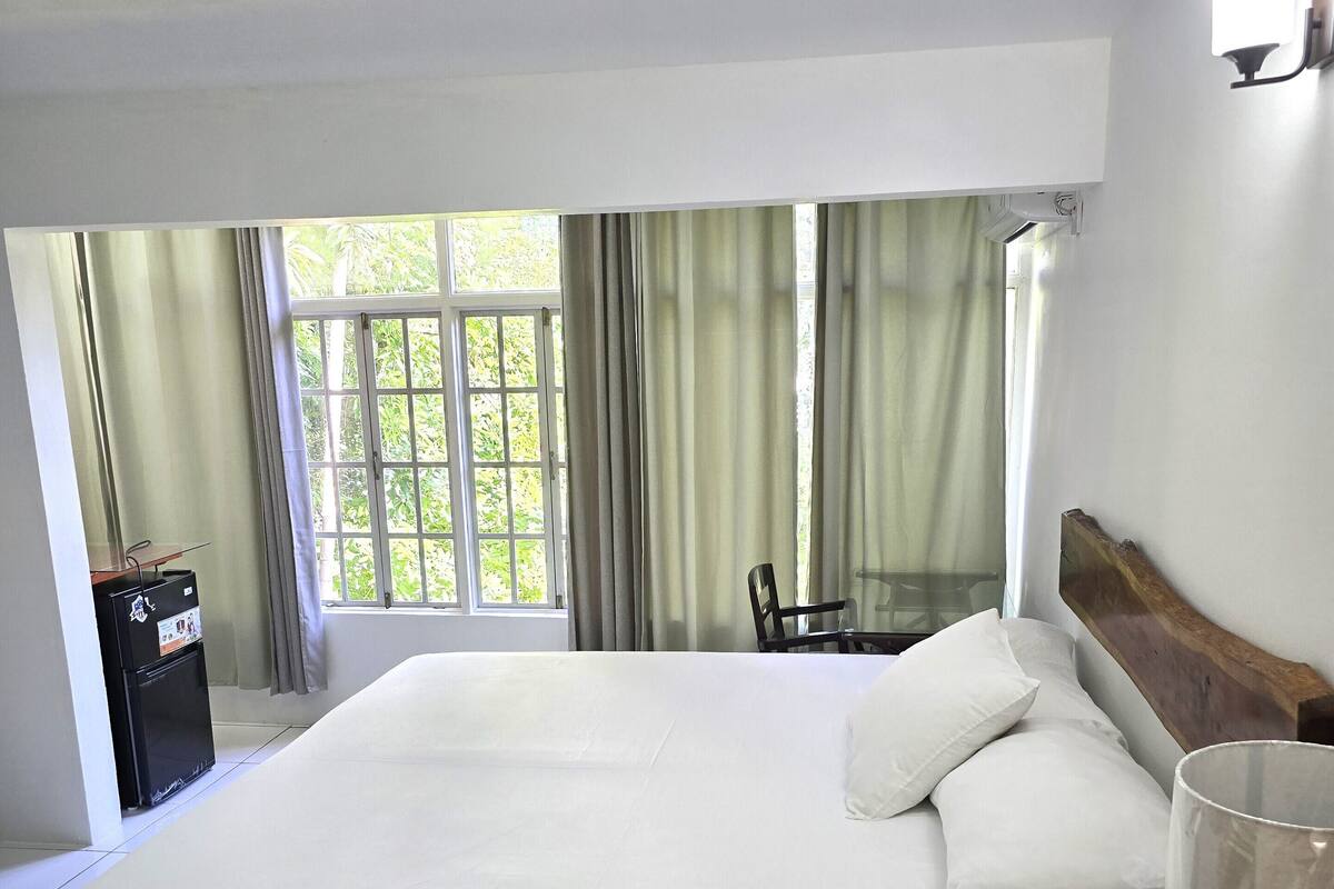A hotel room with a large bed, a small table, a chair, a minibar, and a window with curtains.