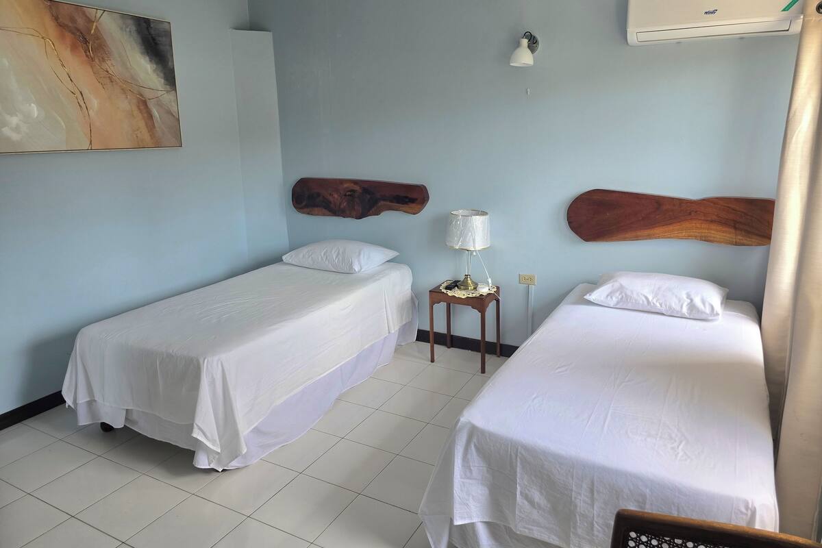 A hotel room with two single beds, a small bedside table with a lamp, a wall-mounted air conditioner, and a painting on the wall.