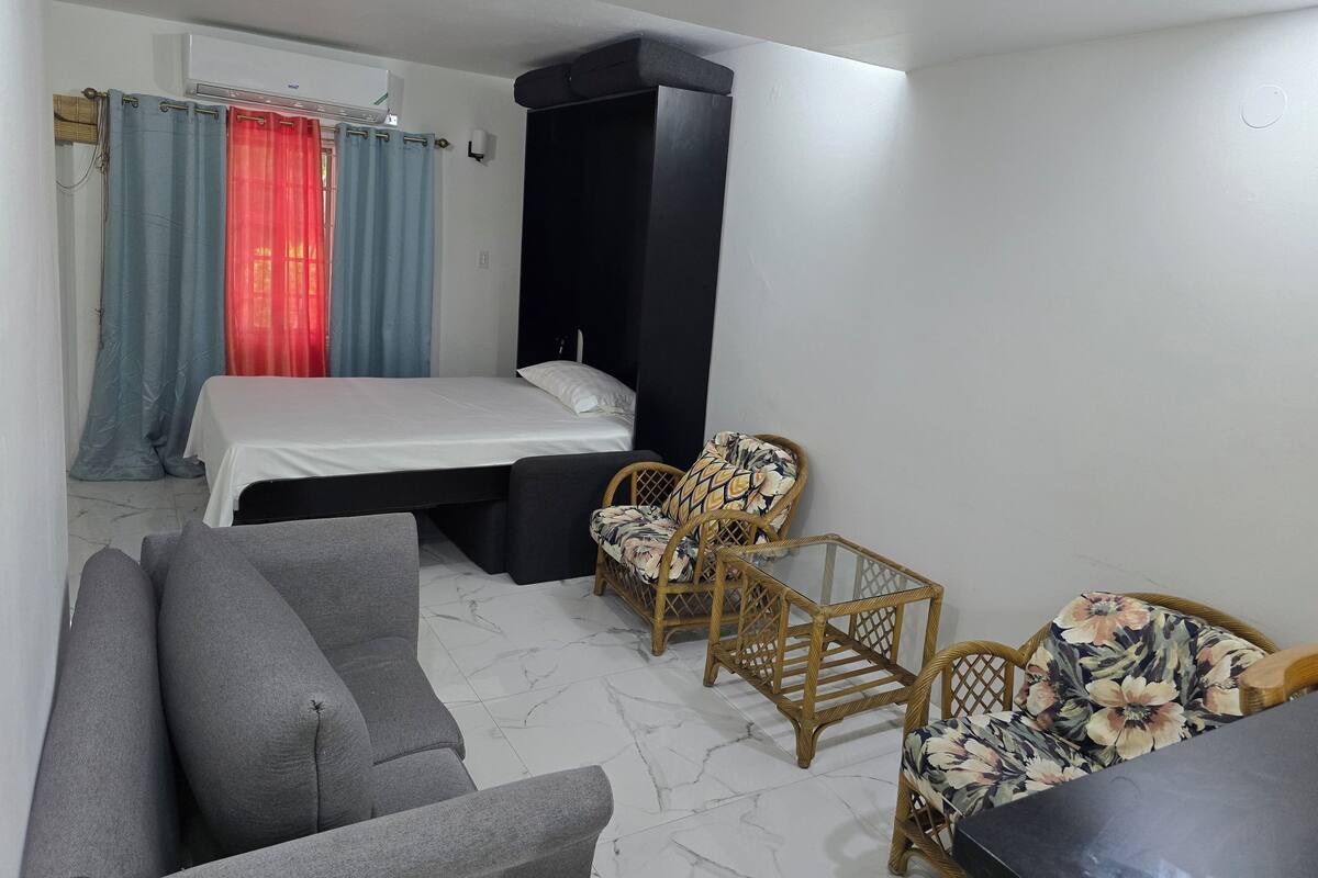 A bedroom with a bed, a grey sofa, two wicker chairs, and a black wardrobe.