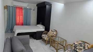 Un dormitorio con cama, un sofá gris, dos sillones de mimbre y un armario negro.