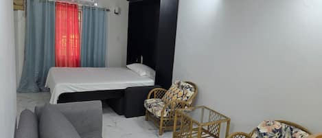 A bedroom with a bed, a grey sofa, two wicker chairs, and a black wardrobe.
