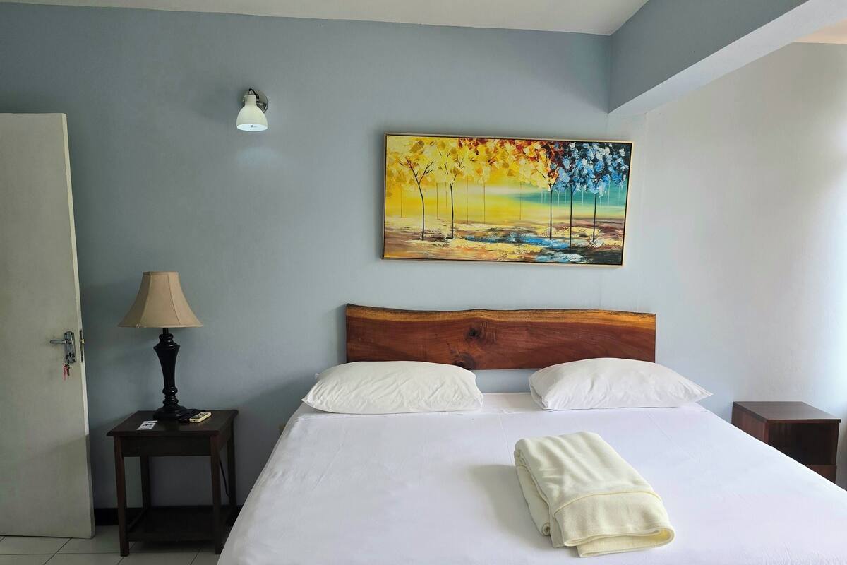A neatly made bed with white linens, a wooden headboard, and a painting of trees on the wall.
