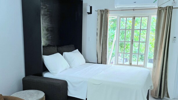 Una habitación de hotel moderna con una cama grande, un cabecero oscuro, una mesita redonda pequeña, un sofá marrón y vistas a zonas verdes por la ventana.