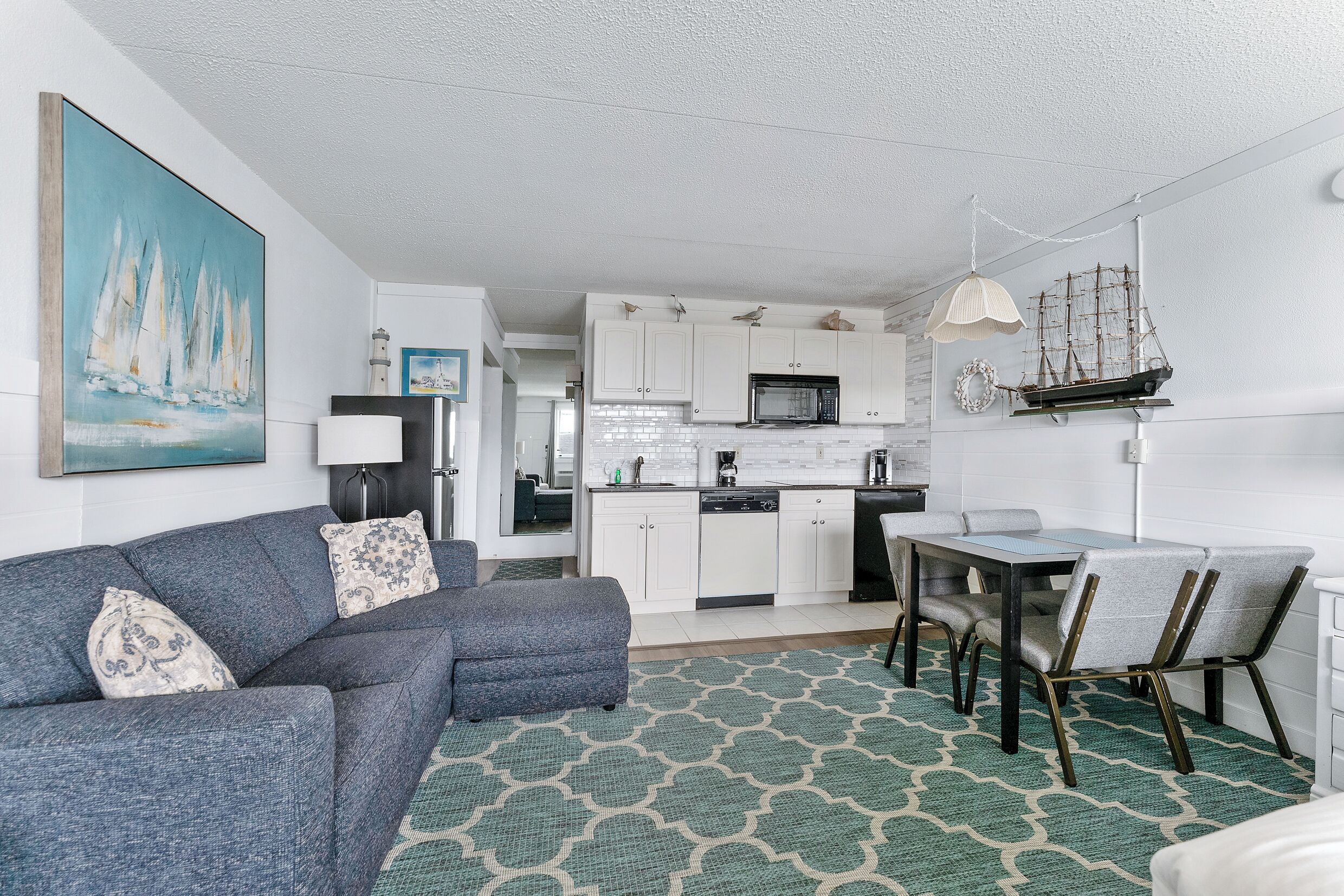 A compact living space with a sofa, dining table, and chairs, a kitchen area with white cabinets and a microwave, and a nautical-themed wall decoration.