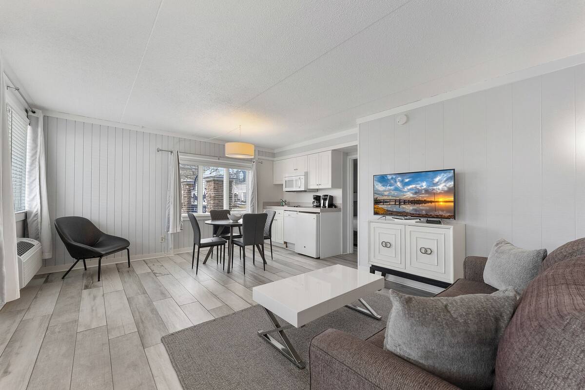 A modern living room with a sofa, a dining area with chairs, a kitchen with white cabinets, and a TV on the wall.