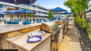 Un espace bar extérieur doté d’un comptoir en pierre, d’un barbecue et de chaises longues placées sous des parasols bleus.