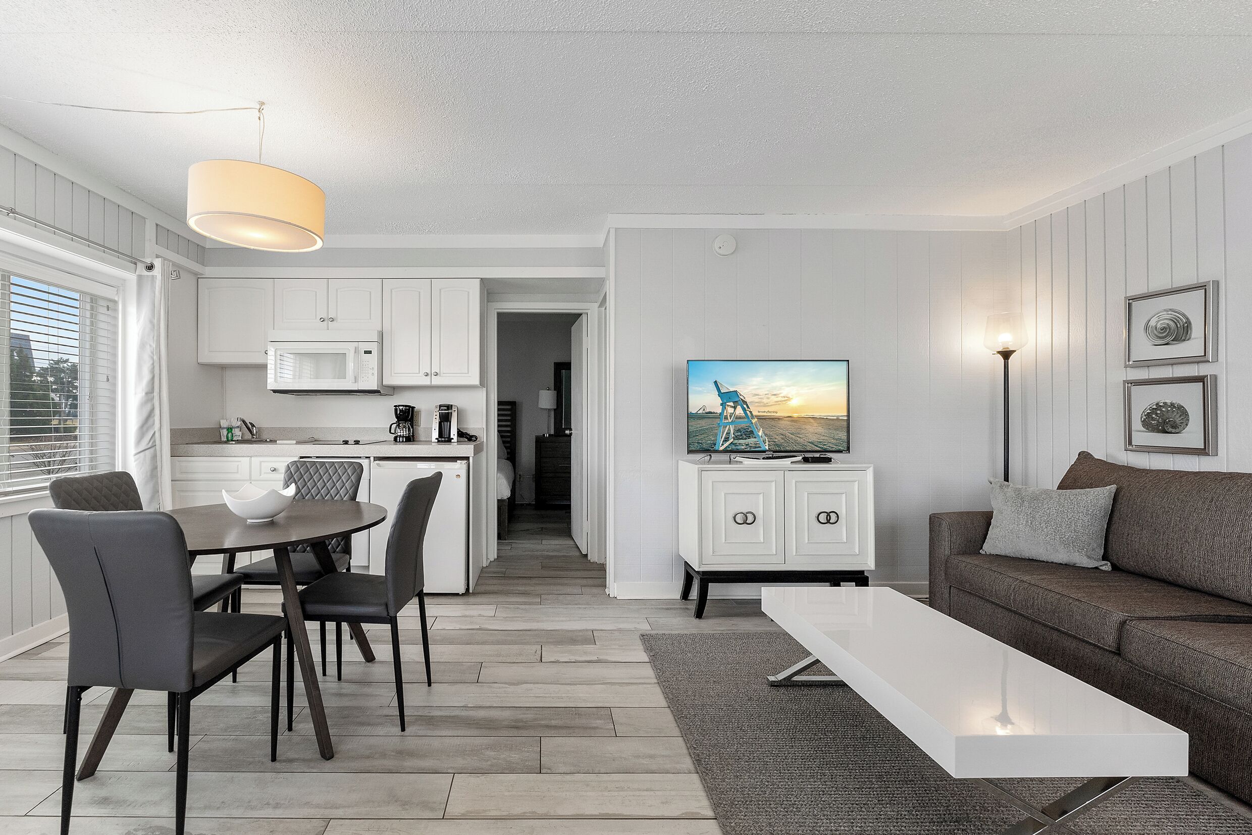 Uma sala de estar moderna com uma televisão de tela plana, uma área de refeições com mesa e cadeiras, e uma cozinha com armários brancos e um micro-ondas.