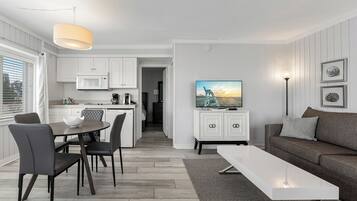 Uma sala de estar moderna com uma televisão de tela plana, uma área de refeições com mesa e cadeiras, e uma cozinha com armários brancos e um micro-ondas.