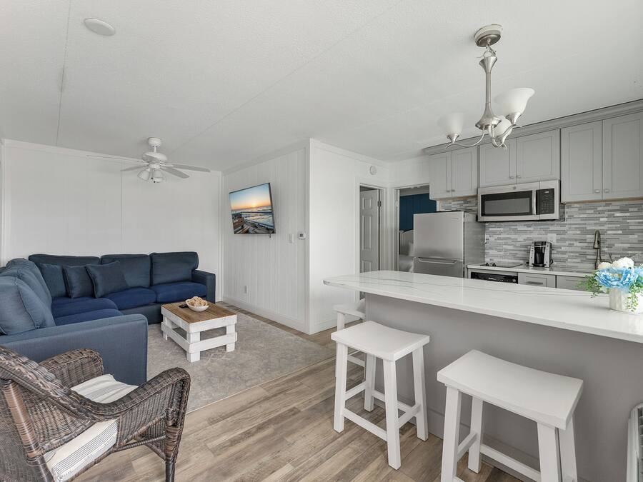 A modern living room with a blue sofa, wicker chairs, and a white coffee table. A kitchen with a microwave and a refrigerator is visible in the background.
