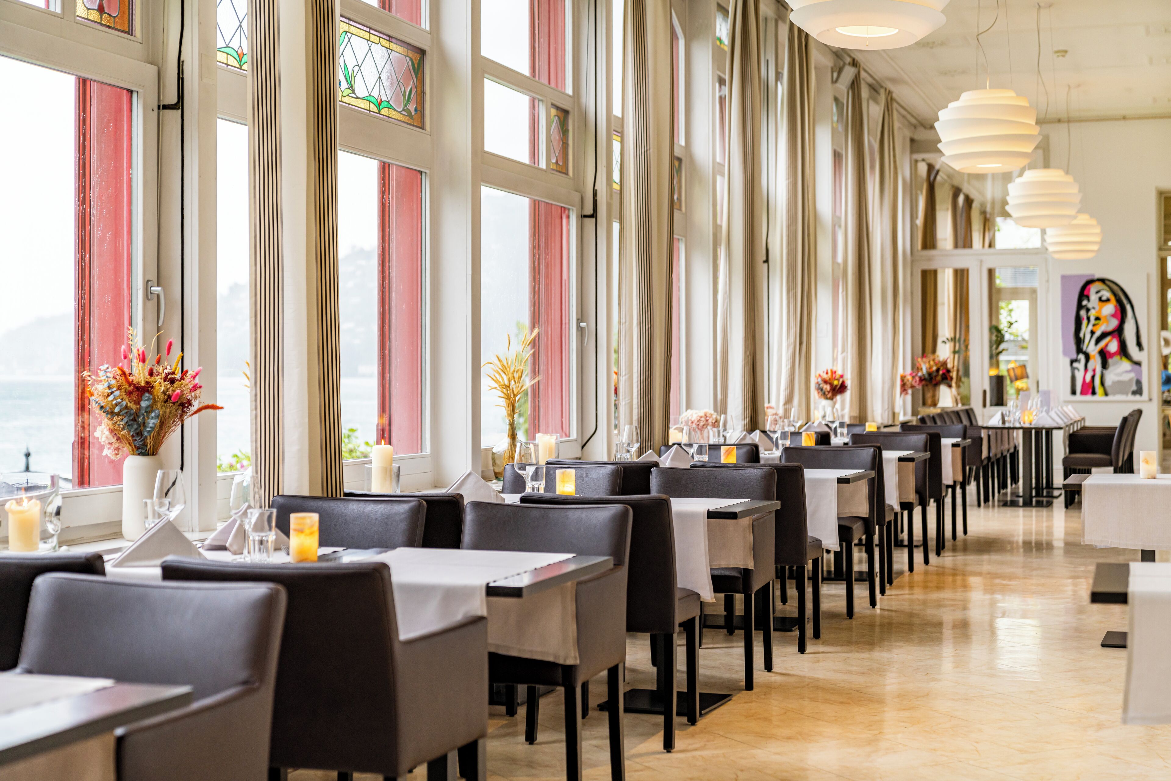 A restaurant with a long table layout, large windows, and modern pendant lighting.