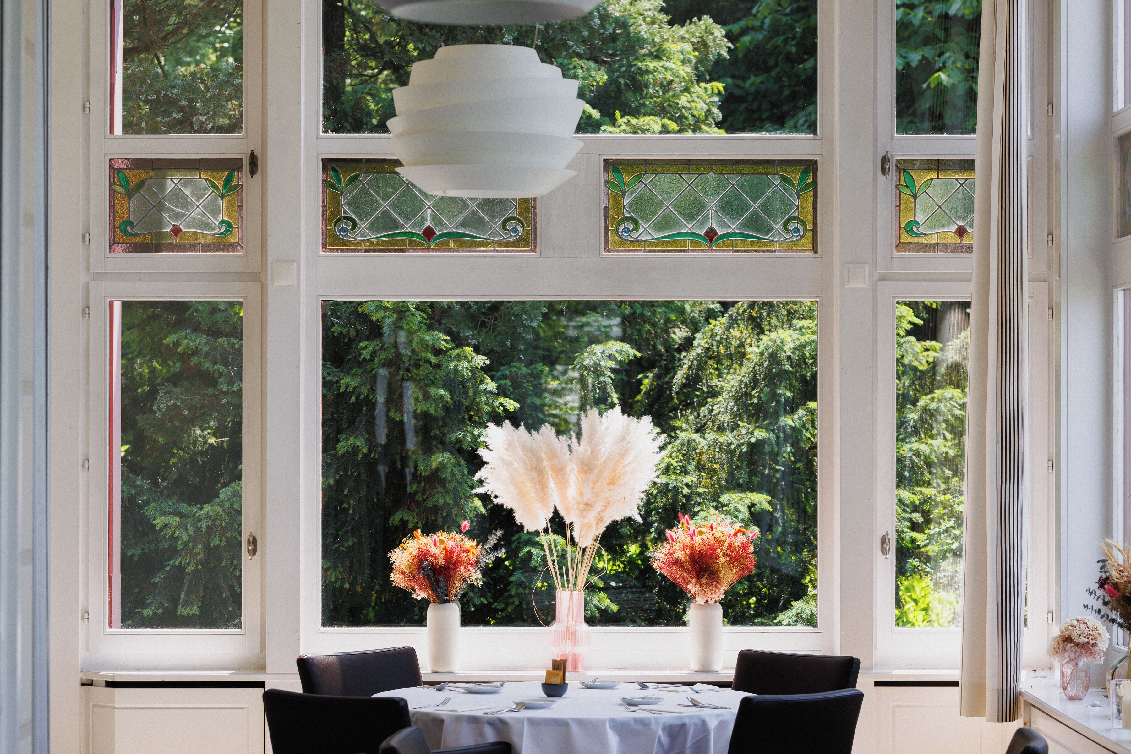 Un coin repas avec une table dressée pour quatre personnes, de grandes fenêtres ornées de vitraux décoratifs et un luminaire suspendu.