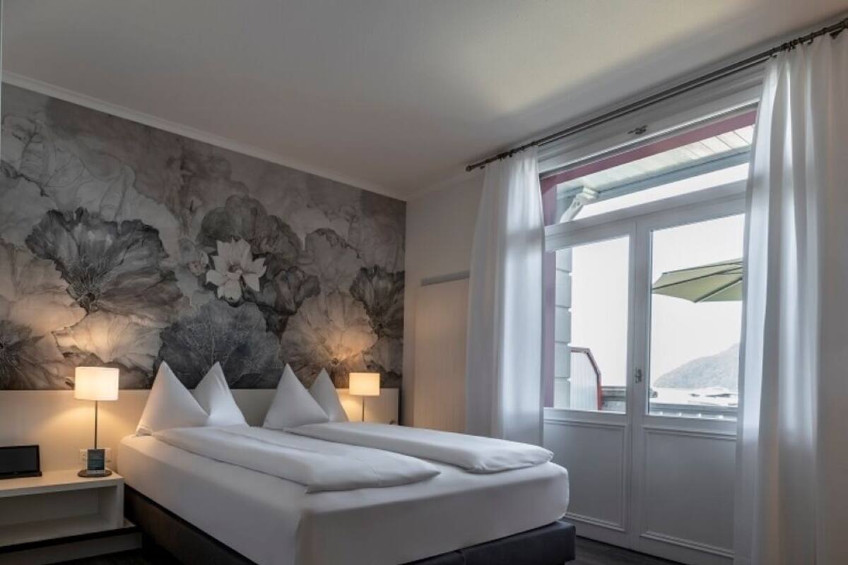 Una habitación de hotel moderna con una cama grande, una mesita de noche con lámpara, un mural de pared de piedra y una ventana con cortinas.