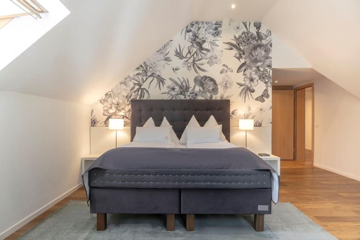 Un dormitorio con una cama grande, un cabecero oscuro, dos mesitas de noche con lámparas y un papel tapiz floral.