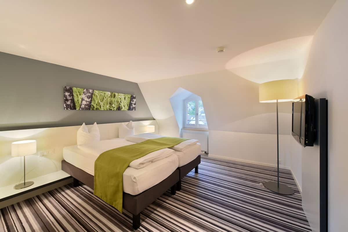 Una habitación de hotel moderna con una cama grande, un televisor de pantalla plana, un escritorio con lámpara y un ventanal con vistas a zonas verdes.