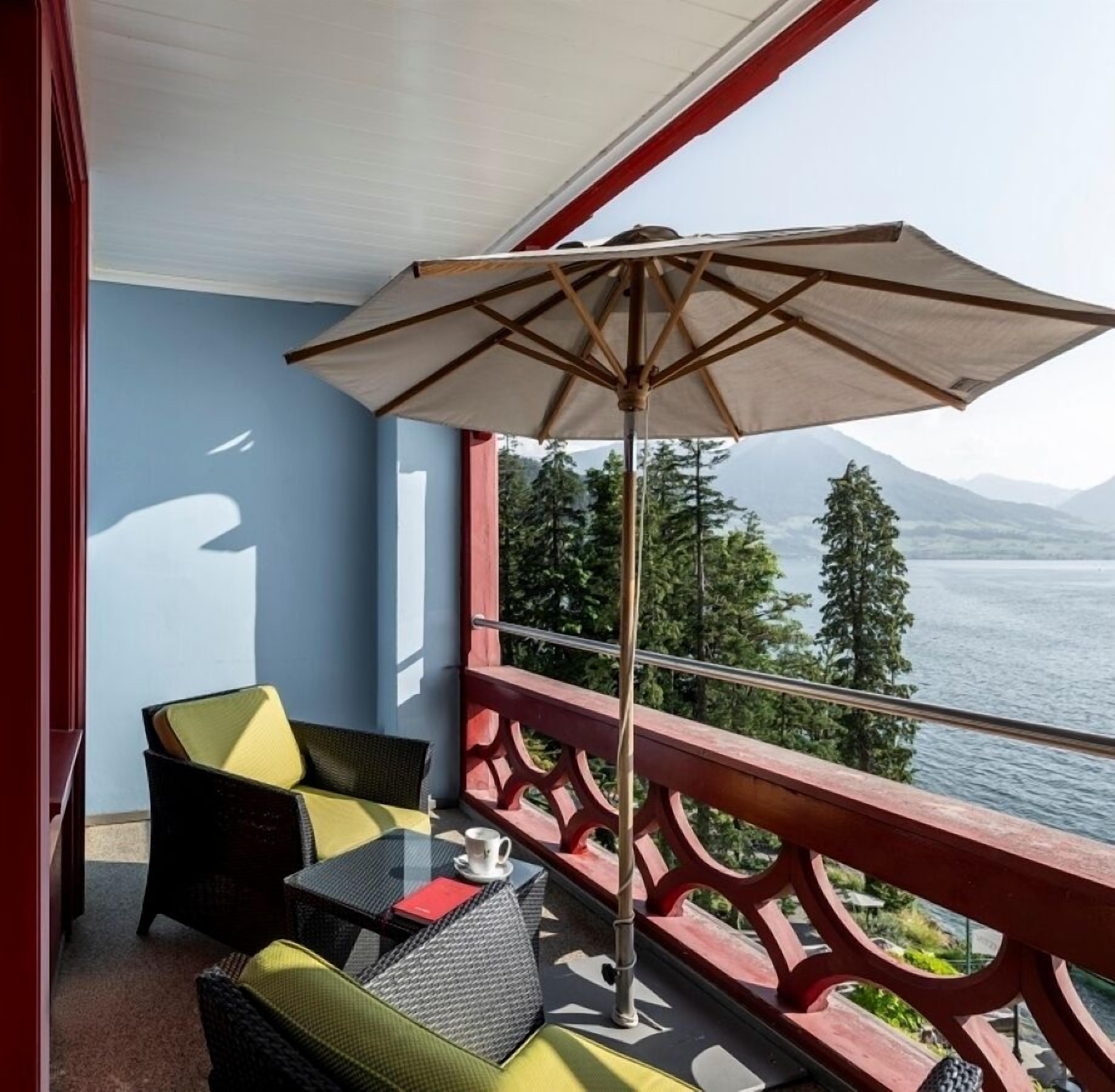 Un balcon avec une table et des chaises, un grand parasol, et une vue sur un lac et des montagnes.