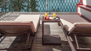 Terraza de madera con sillones de mimbre y una mesa con fruta y jugo, con vista a un cuerpo de agua y montañas.