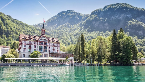 Un edificio junto al lago con una torre, rodeado de árboles y montañas.